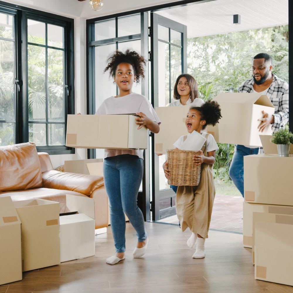 Happy African American young family bought new house. Mom, Dad, and child smiling happy hold cardboard boxes for move object walking into big modern home. New real estate dwelling, loan and mortgage.